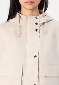 Μπεζ ζακέτα με κουκούλα, με κούμπωμα μπροστά με κουμπιά, τάκους στον κόμπο της κουκούλας και δύο μπροστινές τσέπες. Λείο ύφασμα με μινιμαλιστικό σχέδιο.