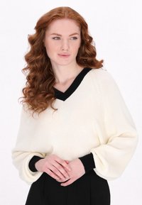 Maglione bianco lavorato a maglia con scollo a V nero e polsini neri, caratterizzato da maniche voluminose, indossato con pantaloni neri. Texture liscia, dettagli di design minimalisti.