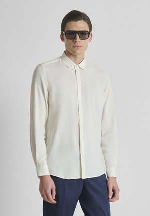 Hombre con camisa blanca abotonada, pantalones azul oscuro y grandes gafas de sol negras, de pie contra un fondo liso y claro.