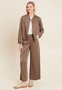 Giacca bomber in satin marrone con bottoni a pressione, abbinata a pantaloni larghi in satin marrone. Il modello indossa una camicia bianca e sneakers beige.