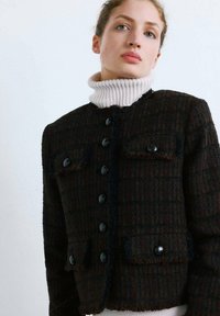 Czarna i brązowa marynarka z tweedu z kremowym, prążkowanym golfem, zapinana na guziki, z dwoma kieszeniami na piersi oraz postrzępionym dołem. Całość zdobi teksturowany wzór w kratę.
