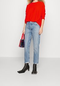 Rotes Langarmoberteil, hellblaue gerade Jeans mit fransigem Saum, schwarze Knöchelstiefel und eine kleine mehrfarbige transparente Handtasche.