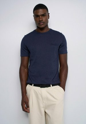 Homme aux cheveux courts portant une chemise bleu marine à manches courtes et un pantalon beige avec une ceinture noire, debout devant un fond clair uni.