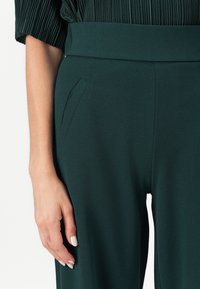 Tamnozelene hlače s širokim pojasom, koje imaju suptilnu teksturu i bočne džepove. Glatka, lepršava tkanina seže do gležnjeva.