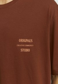 Camiseta de algodón marrón con texto impreso en serigrafía: "COMUNIDAD CREATIVA DE ORIGINALES ESTUDIO" en beige claro. Cuello redondo, mangas cortas.
