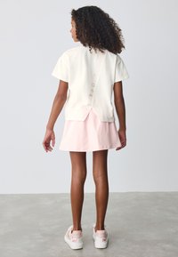 Top crem deschis cu mâneci scurte și decupaj la spate, cu accente roz, asortat cu o fustă roz pal și pantofi sport albi cu detalii florale.