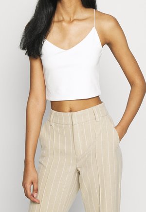 Kvinde iført en hvid cropped tank top og beige højtaljede bukser med hvide lodrette striber, stående med den ene hånd i lommen.
