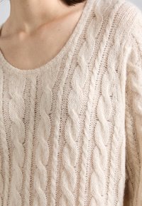 Nära bild av en person som bär en krämfärgad, kabelstickad tröja med rund halsringning, som visar hals och övre bröst.