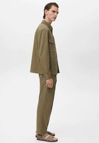 Conjunto de dos piezas en verde oliva que incluye una camisa de botones con un bolsillo en el pecho y pantalones rectos, hechos de una tela texturizada; combinado con sandalias.