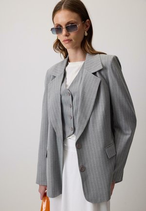 Gray striped blazer with wide lapels, front buttons, and a layered vest beneath. Model holds an orange bag. Textured fabric.