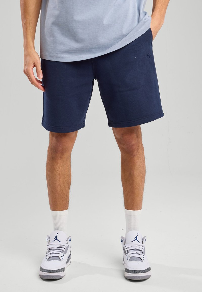 Navy blue sweat shorts made of cotton blend; elastic waistband, side pockets, and a relaxed fit. Paired with white sneakers and white socks.