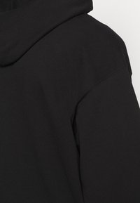 Sweat à capuche noir en tissu doux et texturé avec une coupe décontractée, une capuche bien marquée et des coutures qui créent un design épuré et minimaliste.