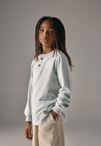 Grijs henleyshirt met lange mouwen en drie knopen aan de voorkant, zachte textuur en een logo op de borst; gecombineerd met lichtbeige broek.