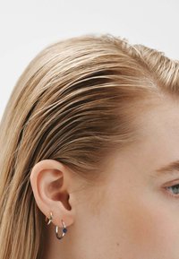 Pendientes de aro de oro y plata en la oreja de una modelo, con un acento verde y una gema azul, que complementan su cabello recto y rubio.