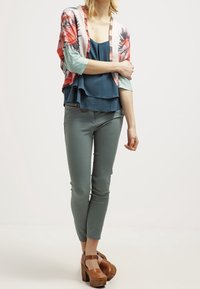 Kimono-Jacke mit floralem Muster und kurzen Ärmeln, übereinander liegendes türkisfarbenes Tanktop, schmale grüne Hose und braune Plateauschuhe.