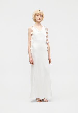 Moteris su trumpais garbanotais šviesiais plaukais dėvi ilgą baltą suknelę be rankovių su permatomais gėlių rašto šonais ir baltus atvirus sandalus.