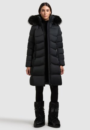 Abrigo tipo puffer negro con capucha de piel sintética, patrón acolchado, diseño de longitud completa y bolsillos laterales con cremallera. Combinado con botas negras aislantes.