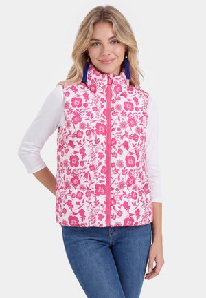 Frau trägt eine weiße Steppweste mit pinkem Blumenmuster, blaue Quastenohrringe, weißes Langarmshirt und blaue Jeans, lächelt mit der Hand in der Hüfte.