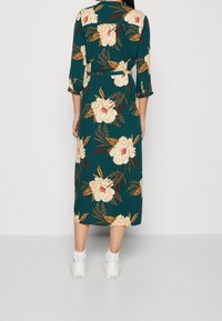 Abito a stampa floreale in teal, con maniche a tre quarti, vita cinchata e lunghezza midi, abbinato a sneaker bianche.