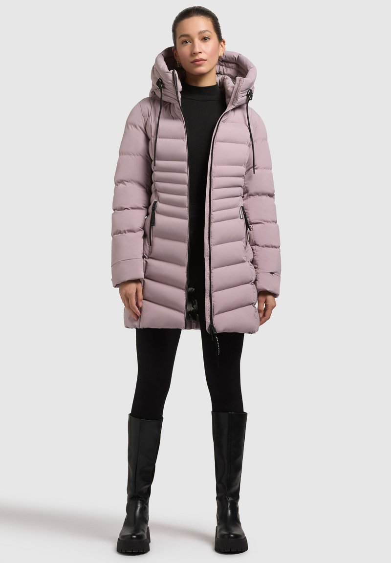 Chaqueta de plumón morado claro con capucha, con segmentos acolchados, bolsillos frontales con cremallera y cordones ajustables. Combinada con leggins negros y botas.