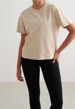 Femme portant un t-shirt beige à manches courtes avec le logo "Aim'n Sportswear" et des leggings noirs côtelés, debout avec une main sur la hanche.