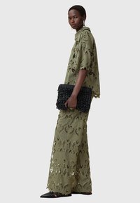 Completo in pizzo verde oliva, composto da una maglietta ampia e pantaloni abbinati con ritagli floreali. Una borsa a tracolla nera con texture e scarpe nere flat completano il look.