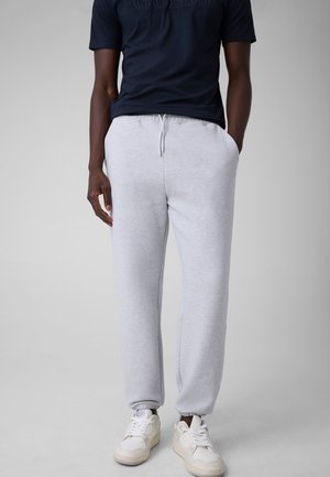 Persona che indossa pantaloni della tuta grigio chiaro con coulisse, scarpe da ginnastica bianche e una maglietta scura a maniche corte, in piedi con le mani in tasca.