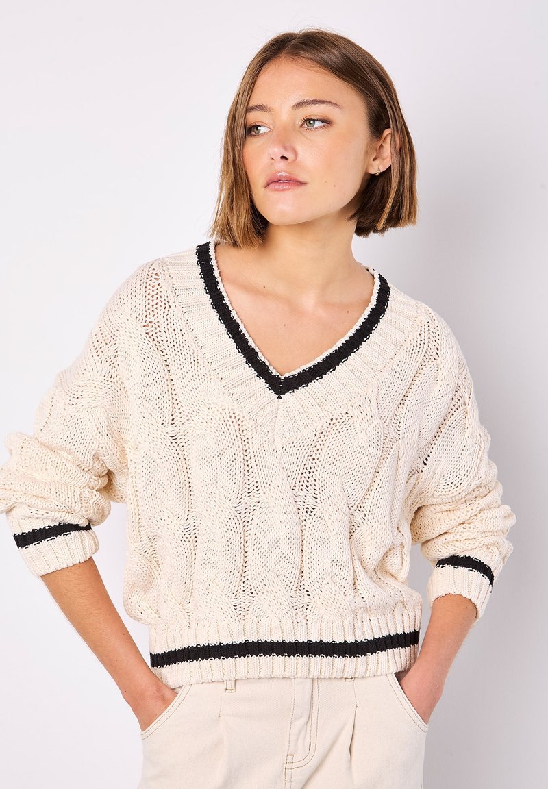 Cremfarbener Strickpullover mit V-Ausschnitt, der ein strukturiertes Zopfmuster und schwarze Streifenakzente an den Bündchen und am Saum aufweist.