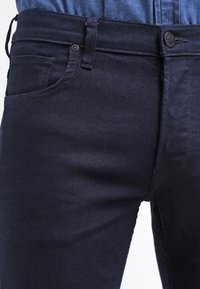 Tummansinisiä farkkuja, joissa on kapea malli, viisi taskua ja nappi suljenta. Rakenteellinen kangas, jossa näkyvät saumat ja hienovarainen kiilto.