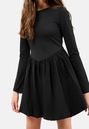 Femme portant une robe noire à manches longues avec un haut côtelé ajusté et une jupe froncée évasée, debout devant un fond uni.