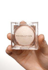 Revolution highlighter i fyrkantig transparent kompakt, med ljus beige puder med shimmer. Visad i en hand mot en vit bakgrund.