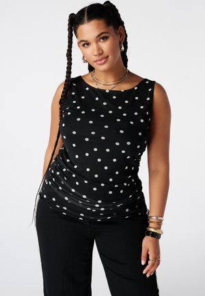 Vrouw met lang gevlochten haar, gekleed in een mouwloze zwarte top met witte stippen en een zwarte broek, poseert tegen een effen achtergrond.