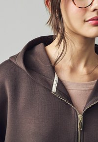Brun zip-up hættetrøje med en blød tekstur, der har en stor hætte og en sølvfarvet lynlås. En lys pink ribbet trøje er synlig under.