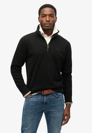Zwarte zip-up sweatshirt met een opstaande kraag en grijze voering, gedragen met een beige shirt eronder, blauwe jeans en een bruine riem.