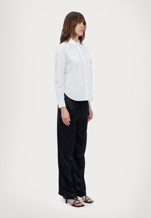 Camisa blanca de manga larga con cuello y un bolsillo frontal, combinada con pantalones de satén negros de pierna ancha y tacones negros con tiras.