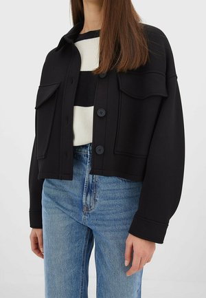 Vrouw draagt een zwart vest met knopen en grote zakken over een zwart-wit gestreepte trui en een blauwe spijkerbroek.