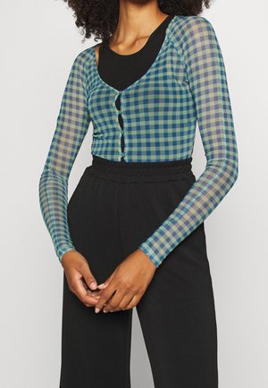 Frau trägt ein langärmliges, grün-blau kariertes Netzoberteil über einem schwarzen Tanktop, kombiniert mit hoch taillierten schwarzen Hosen.