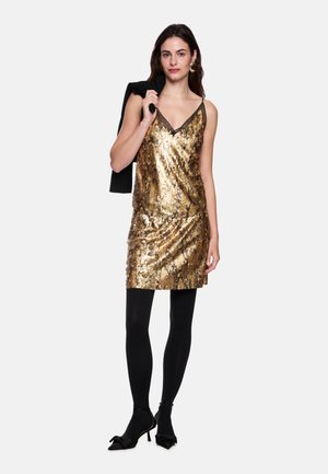 Woman in gold sequin sleeveless dress, black tights, heels, carrying black jacket over shoulder, standing against white background.