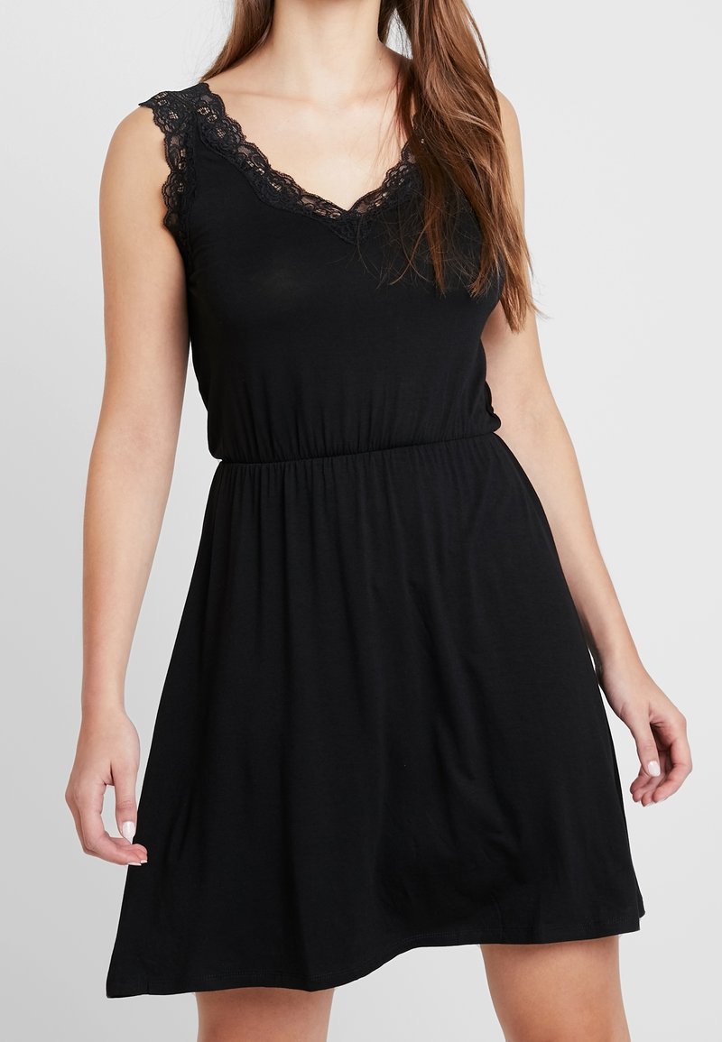 Femme portant une robe noire sans manches avec un bord en dentelle au niveau de l'encolure et une taille élastique, debout devant un fond uni.