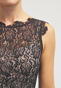 Haut en dentelle noire avec motif floral et bordure festonnée. Texture transparente avec une coupe ajustée, mettant en valeur le décolleté et les épaules.