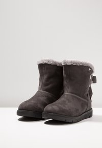 Botas de tobillo de ante gris con un suave forro de pelo sintético gris, suela plana negra y un lazo decorativo en la parte trasera.