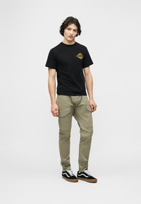 Νεαρός άνδρας που φοράει μαύρο γραφικό μπλουζάκι, μπεζ παντελόνι cargo και μαύρα αθλητικά παπούτσια χαμηλής κοπής με λευκά κορδόνια, πάνω σε απλό φόντο.