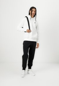 Felpa bianca con cappuccio e un piccolo logo nero, abbinata a pantaloni jogger neri e sneakers bianche. Il modello porta una borsa a tracolla nera.