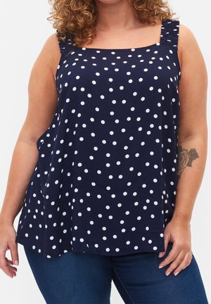 Curly-haired person wearing a navy blue sleeveless top with white polka dots and blue jeans, showing a rose tattoo on the arm.