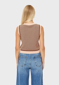 Ärmelloses Strickoberteil in Braun und Hellrosa, mit geometrischem Muster und geripptem Saum. Kombiniert mit hoch geschnittenen blauen Jeans.