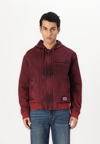Rød zip-up hættetrøje med en tekstureret finish og snor i hætten, med en frontlomme og ribbestrikkede manchetter, parret med blå jeans.