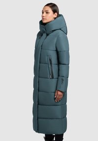 Płaszcz puchowy w kolorze zielonkawo-niebieskim z wysokim kołnierzem, pionowymi kieszeniami na zamek oraz poziomym pikowanym wzorem. Miękka tekstura, długość do kolan i zapięcie na zamek.