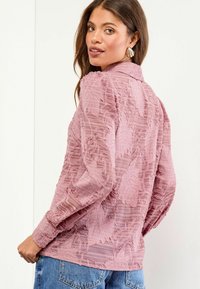 Sheer pink blouse with textured floral embroidery, long sleeves, and collared neck. Features a relaxed fit with a slightly cropped hem.