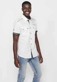 Camisa blanca de manga corta con botones y bolsillos en el pecho, puños en contraste en azul marino y detalles de costura decorativa. Combinada con pantalones de jeans claros.