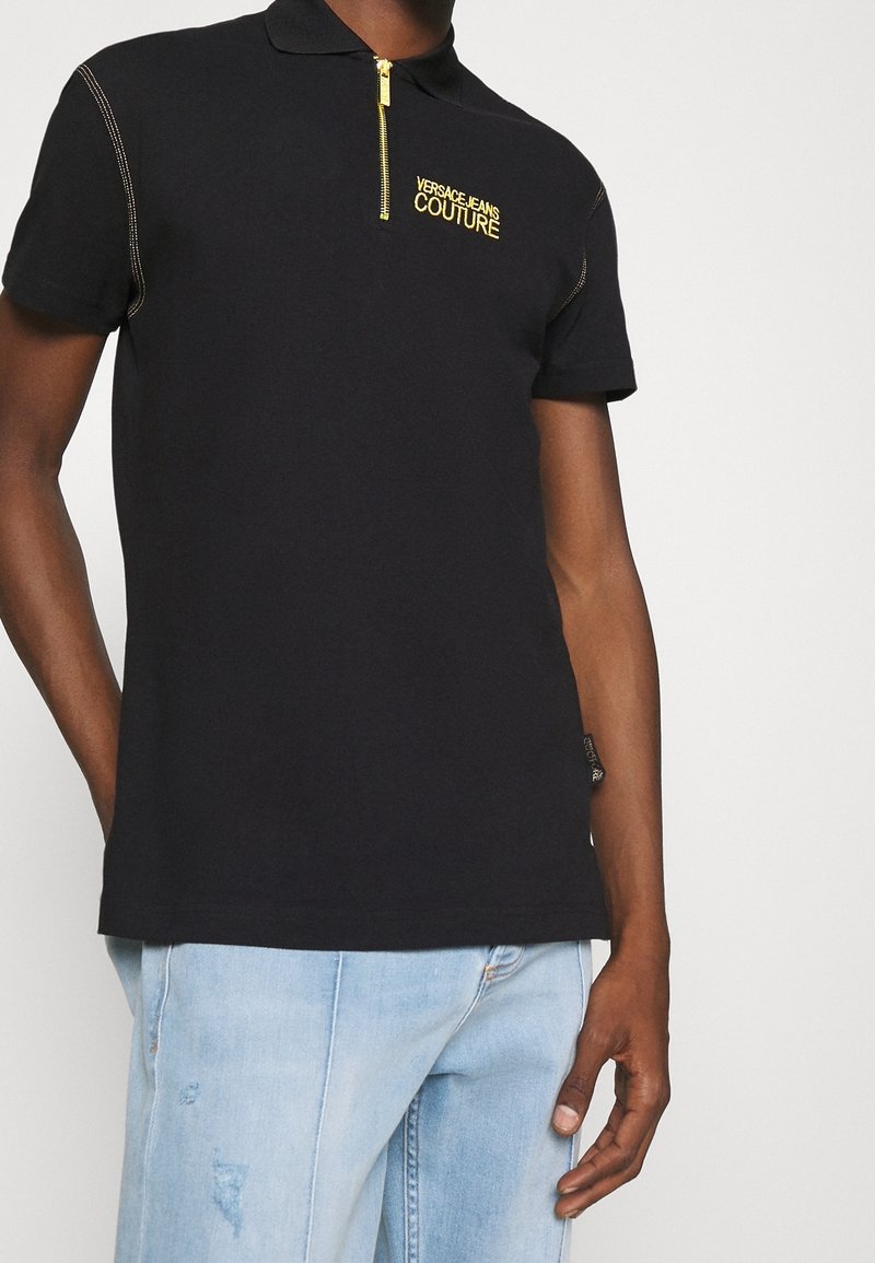 Polo noir à manches courtes avec fermeture éclair dorée et logo "Versace Jeans Couture" sur la poitrine, porté avec un jean en denim bleu clair.